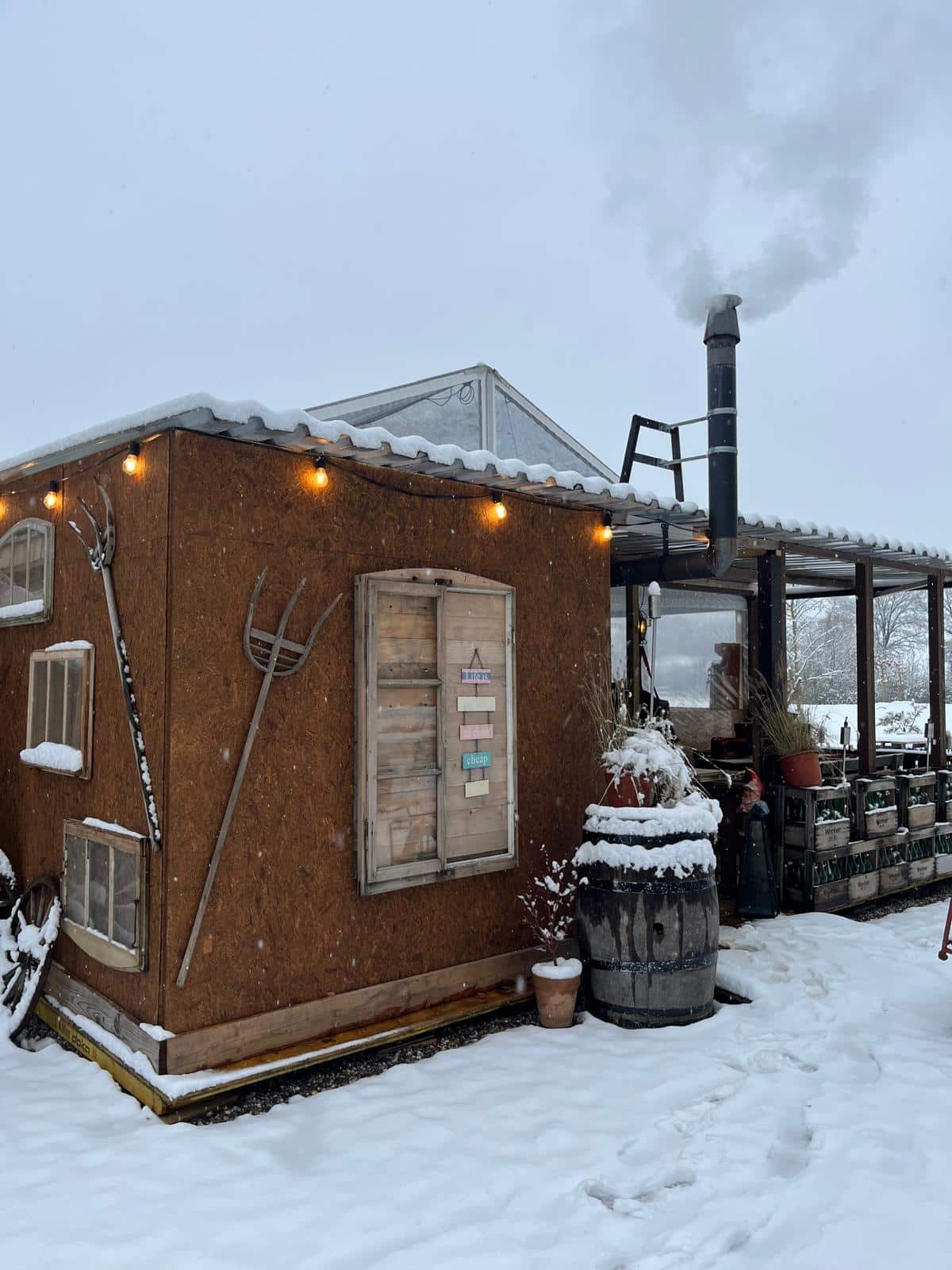Kleines Holzhaus im Schnee mit winterlicher Dekoration und rauchendem Kamin, umgeben von ländlicher Landschaft.